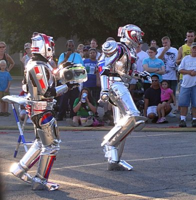 [Two performers in metallic costumes dancing to the music with a crowd in the background.]